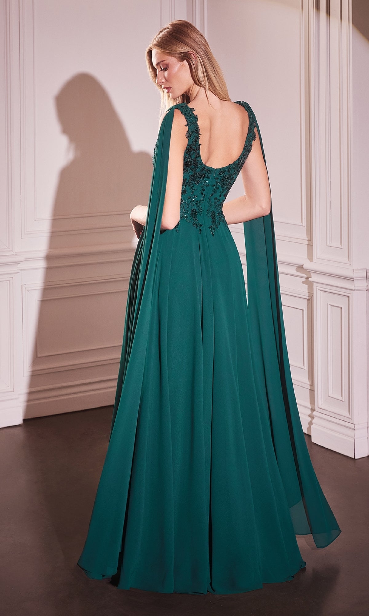 A woman stands in an elegant room with white paneled walls, wearing the Ladivine CDS509 Shoulder Sash Long Formal Dress in dark green, featuring cape sleeves and floral embroidery.
